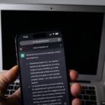 A hand holding a smartphone displaying ChatGPT in front of a laptop 